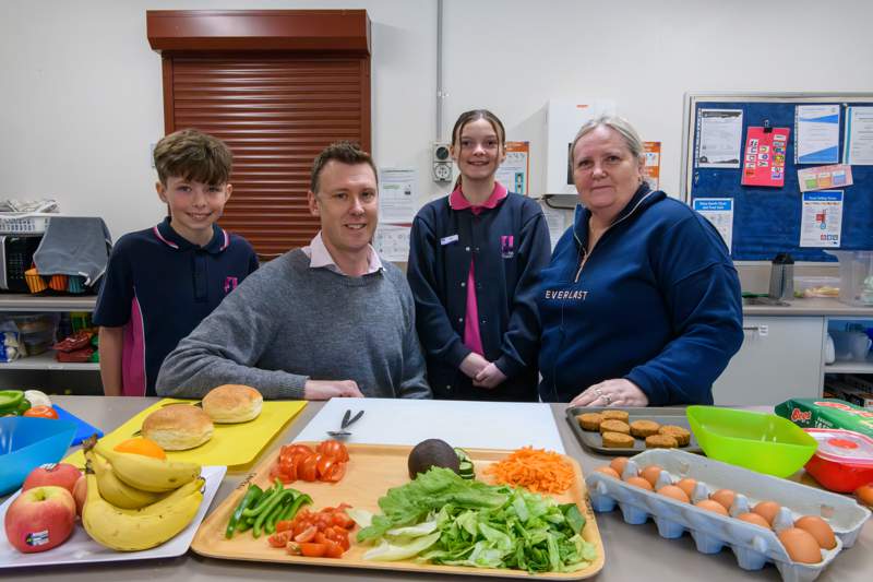 Four people in a school kitchen stand behind a bench with fresh ingredients, including lettuce, tomato, capsicum, carrot, avocado and eggs