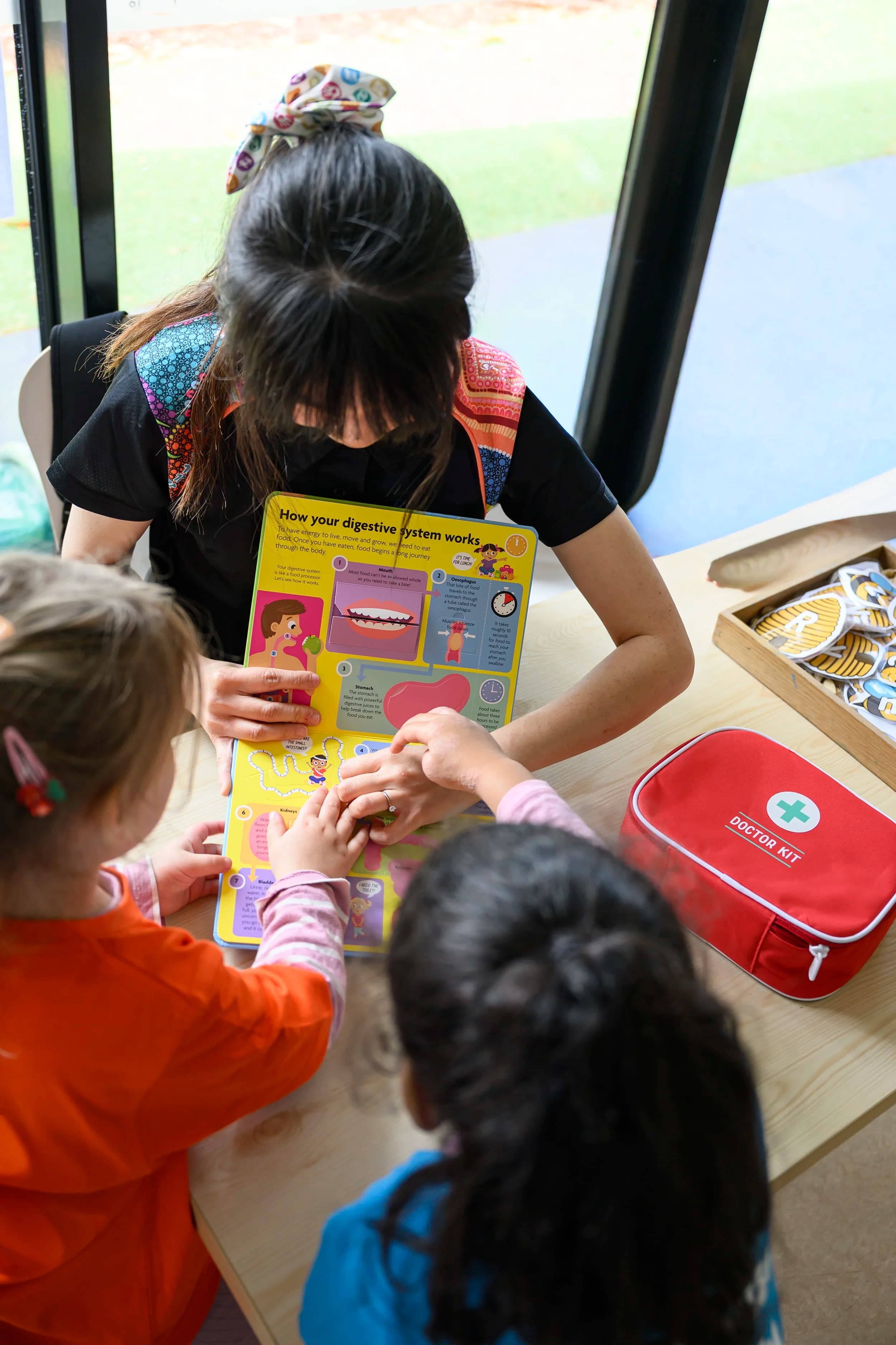 teacher in black shirt showing a colourful book on the digestive system to two children